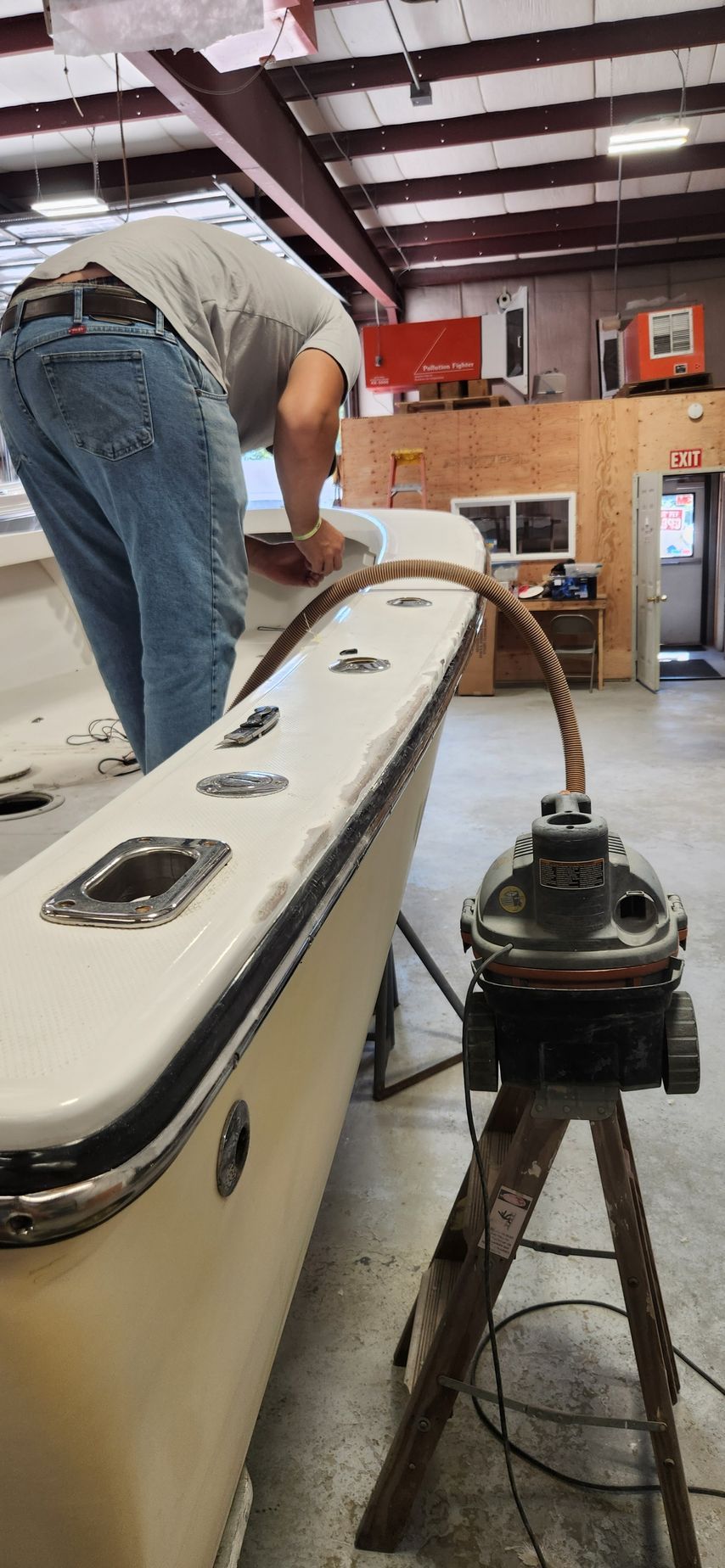 Person works on a boat, connected to a machine via hose. Indoors, industrial setting, white boat, tan wall.