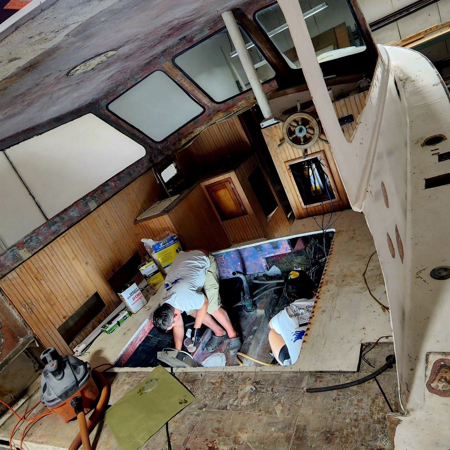 Person working on boat engine, in a boat's interior. Brown and white tones, with tools and wooden panels visible.