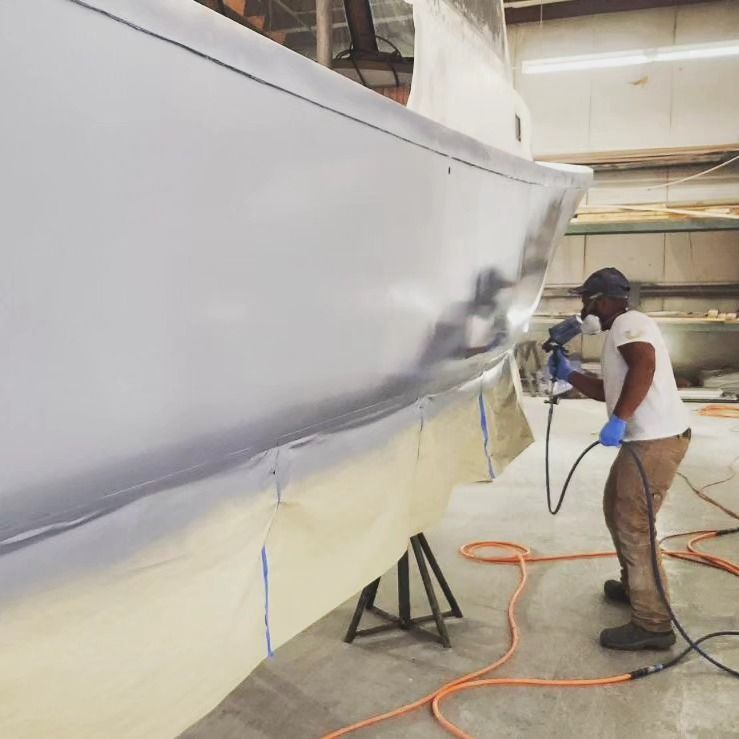 Man in respirator spraying boat hull in a workshop; grey paint, blue tape.