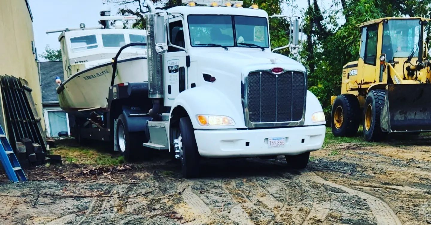 White semi-truck transporting a boat, parked next to a yellow loader.