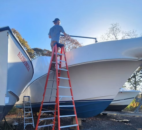 Person on a ladder working on the bow of a white boat, another boat beside it, outdoors on a sunny day.