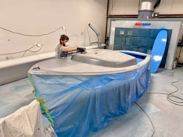 A person sands a gray boat in a workshop. The boat is covered in blue plastic.