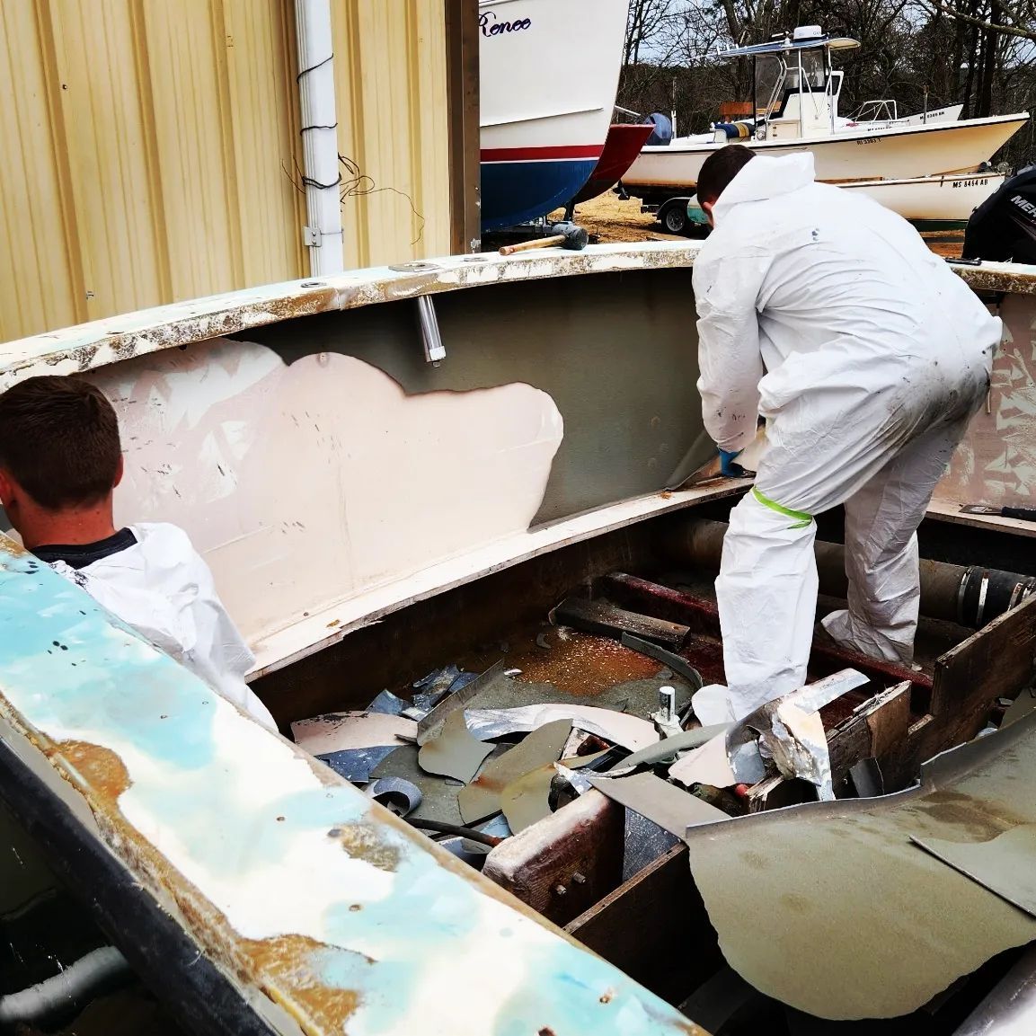 Two people in protective suits demolishing the interior of a boat outdoors.