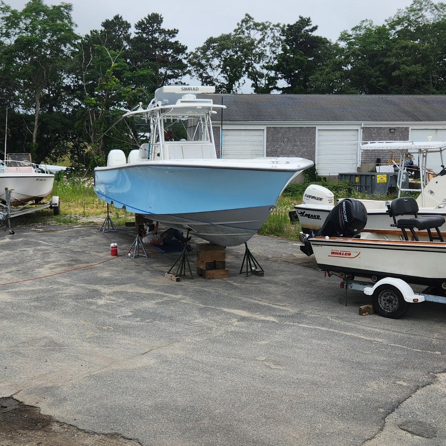 A boat on stands with a light blue hull in a boatyard.  Other boats and a building are visible in the background.