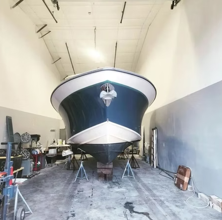 Boat on stands in a gray workshop, blue hull, white trim, and a high ceiling.