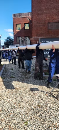 People carry a large white cylindrical object outside a brick building on a gravel path.