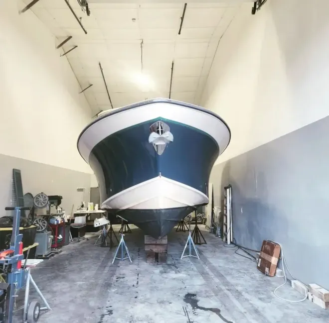 Boat on stands inside a workshop. Blue and white hull, gray concrete floor.
