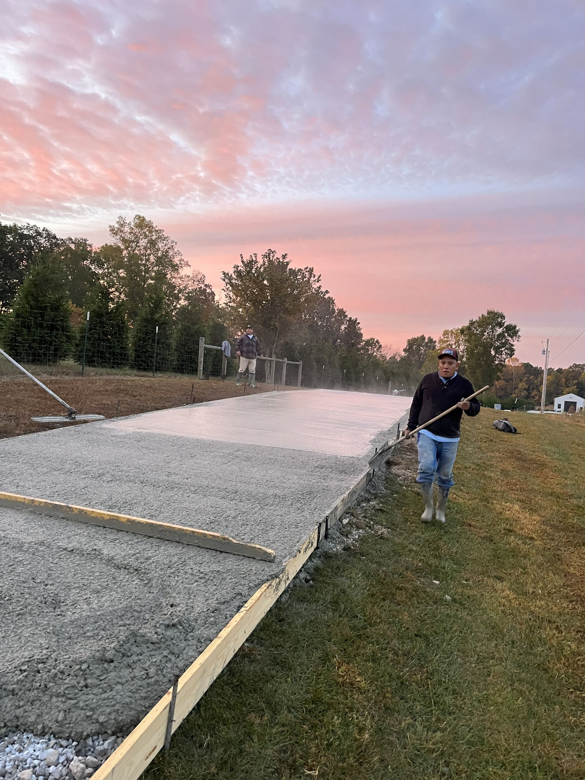 Worker spraying wet concrete in a driveway at sunset, with trees and pink clouds in the background.