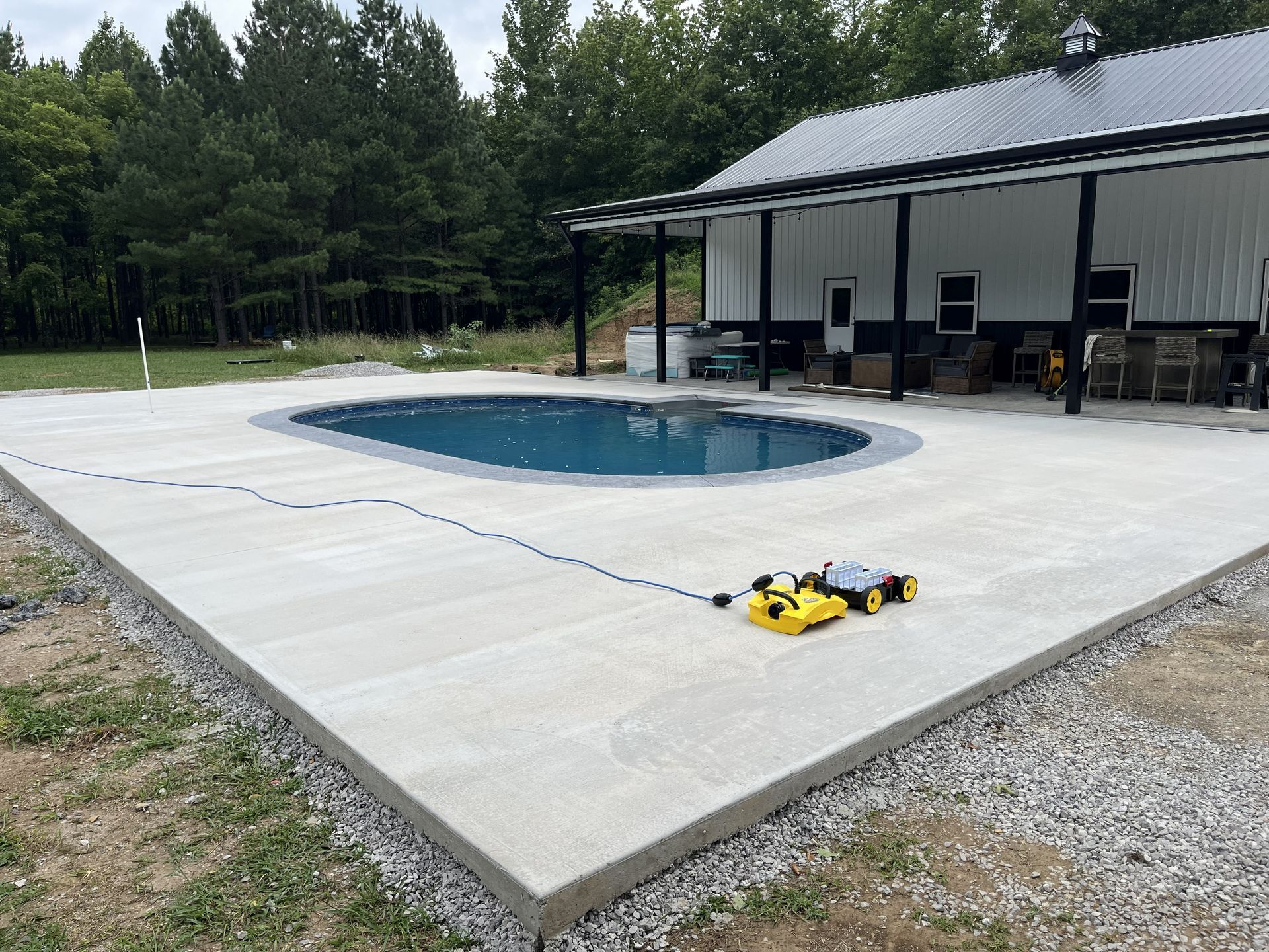 Concrete pool deck with a small oval pool beside a covered patio and wooded yard.