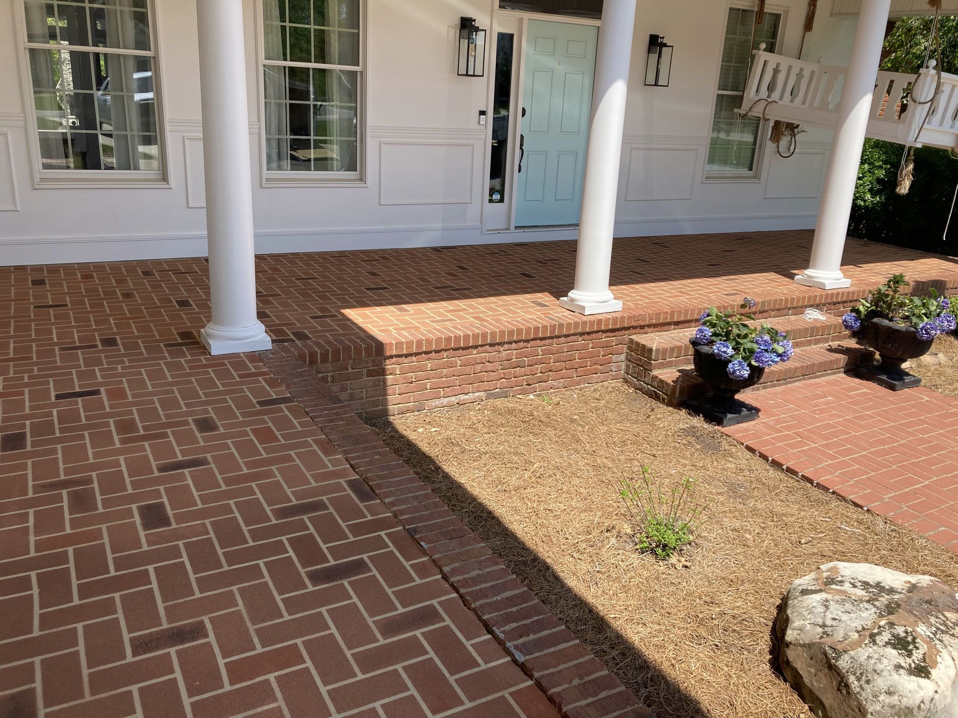 Front porch with white columns, brick walkway, light blue door, and small flower beds with mulch and rocks