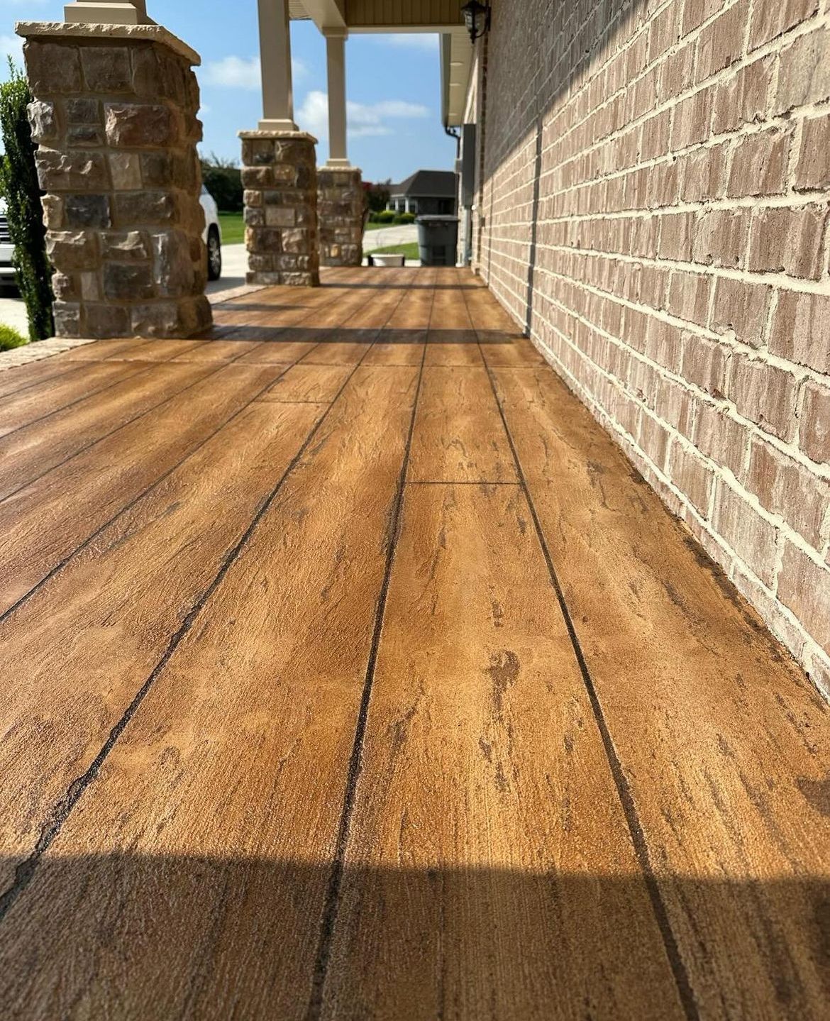 Sunny wooden walkway beside a brick wall and stone columns, leading to an open outdoor area.