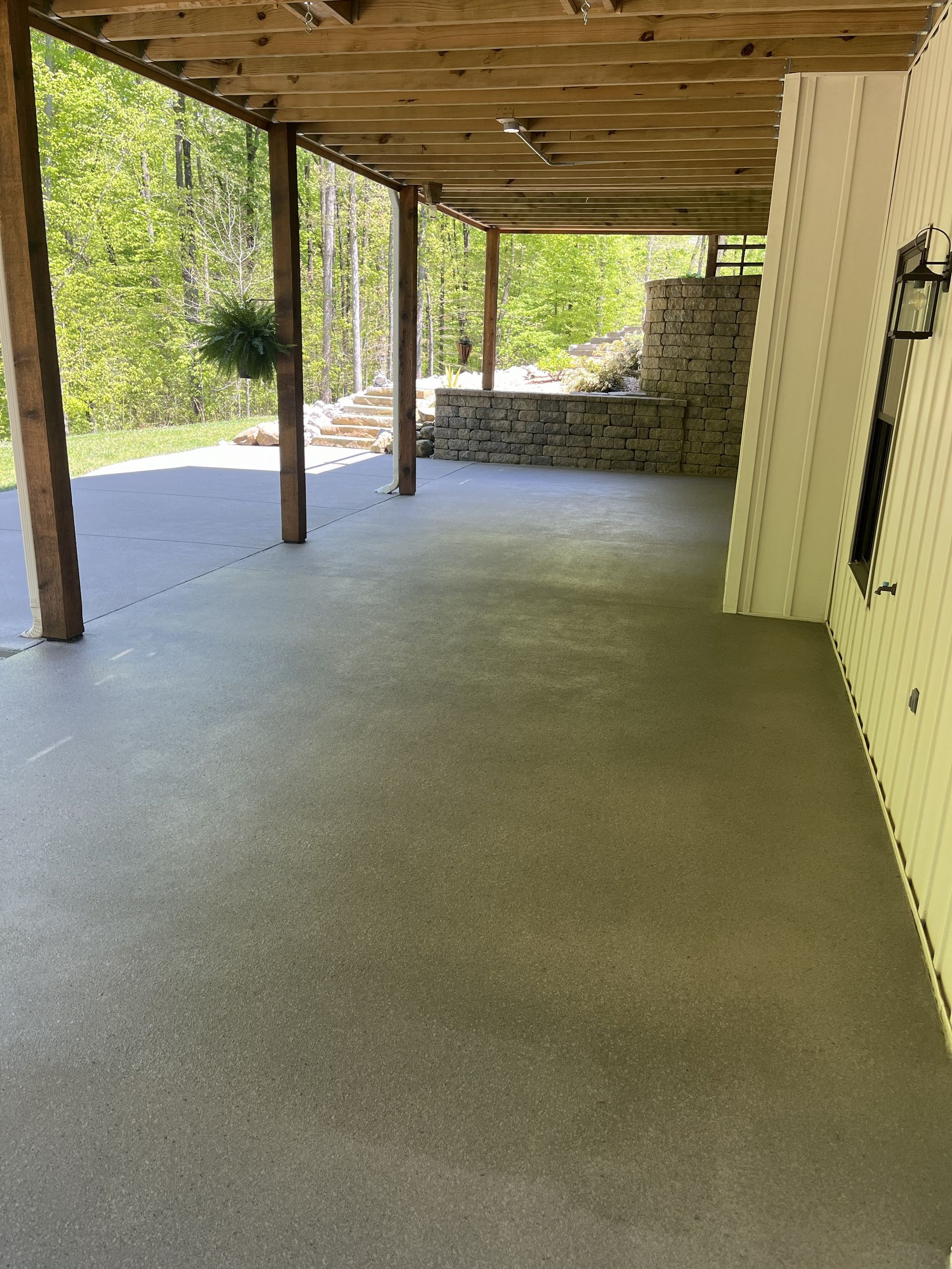 Covered concrete patio with wooden posts and ceiling, beside a stone retaining wall and wooded yard.