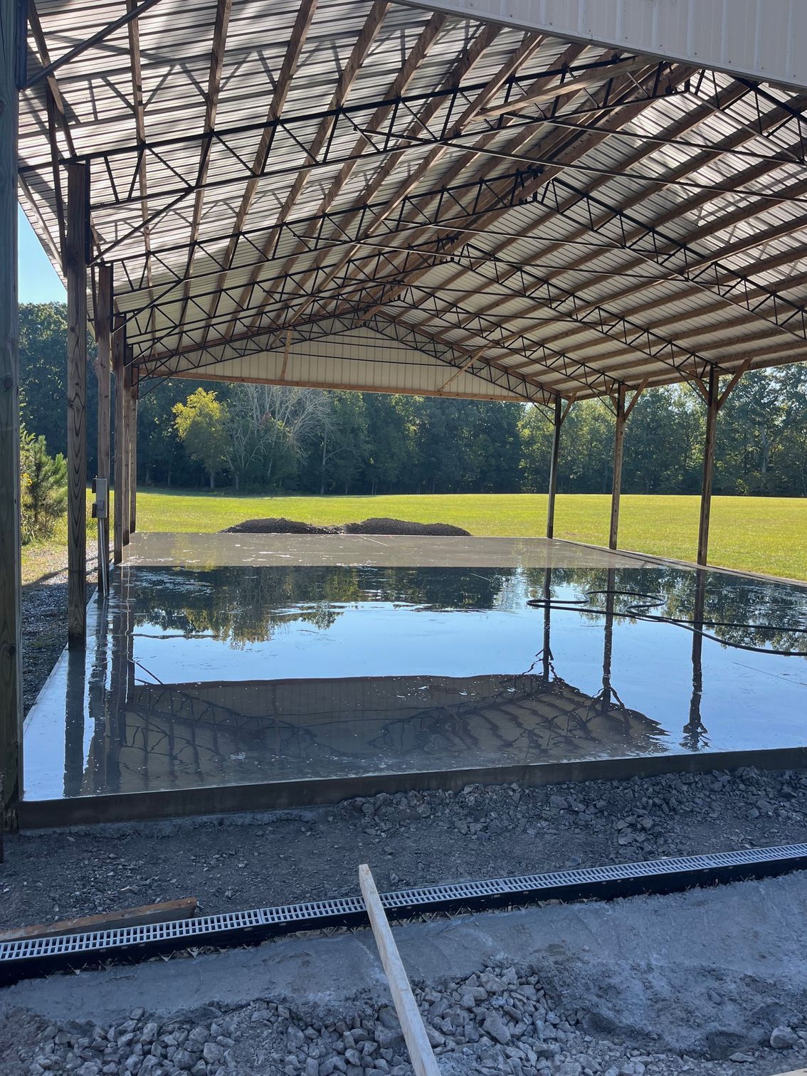 Covered pavilion with a metal roof reflected in a flooded floor, surrounded by grass and trees.