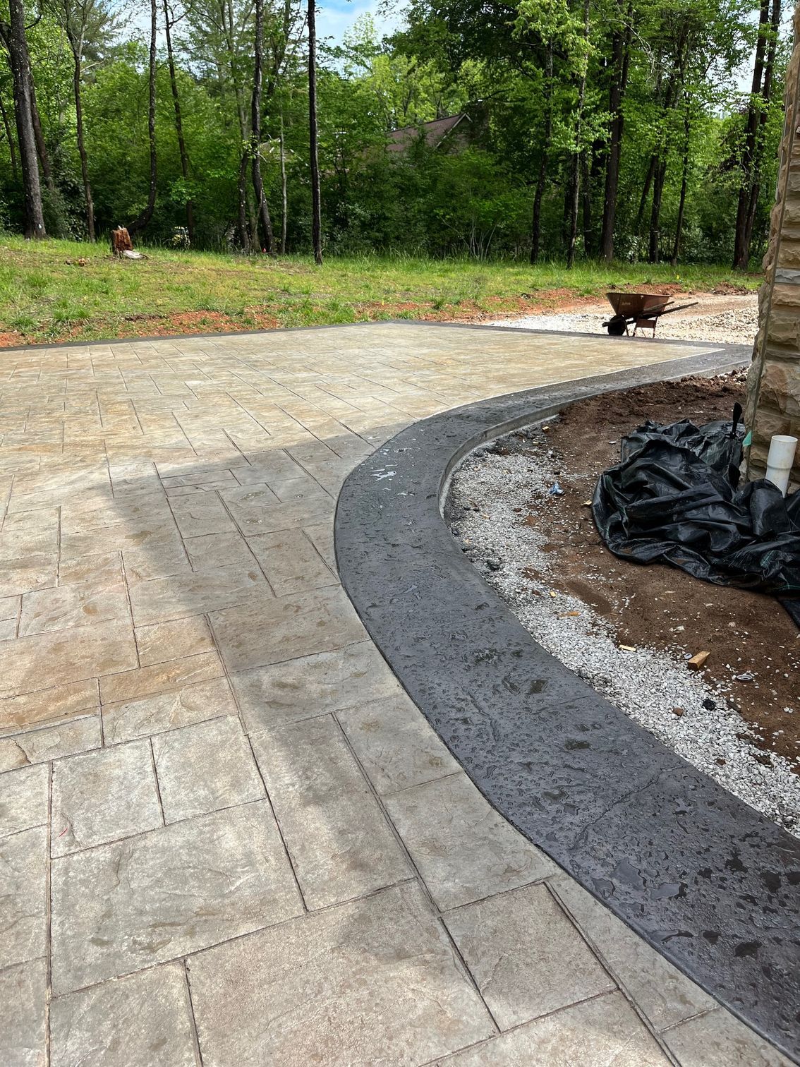 Curved stone patio beside a landscaped yard, with a small dog on the path and trees in the background