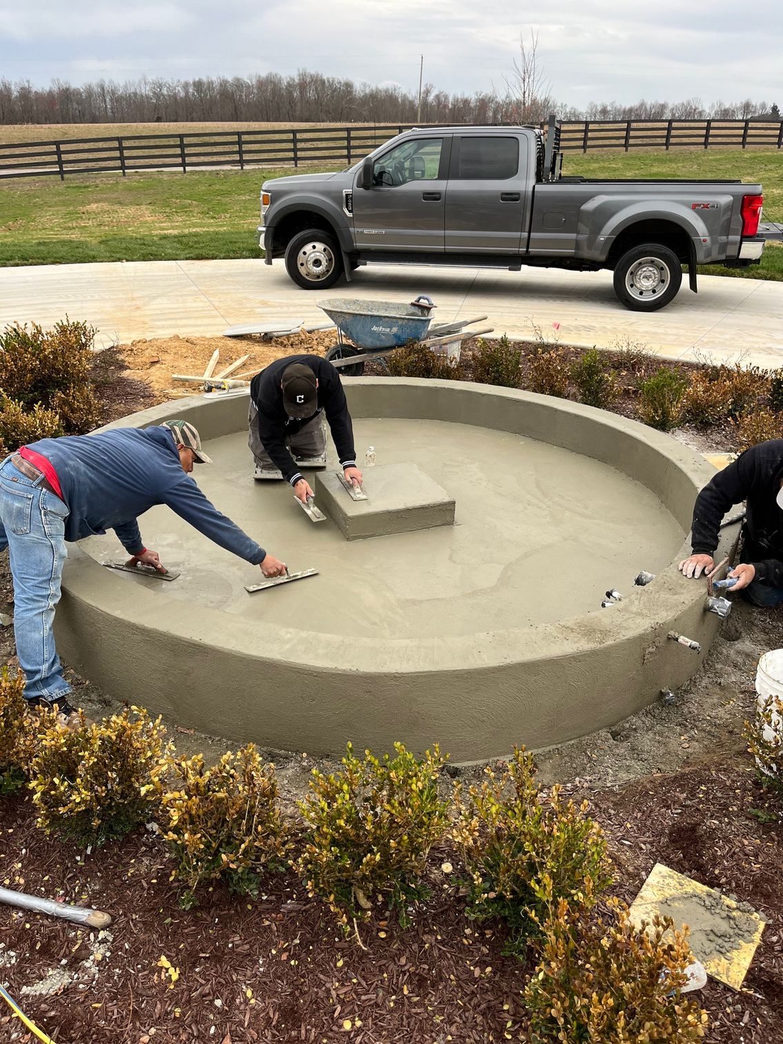Workers smoothing wet concrete in a circular fountain basin near a parked pickup truck.