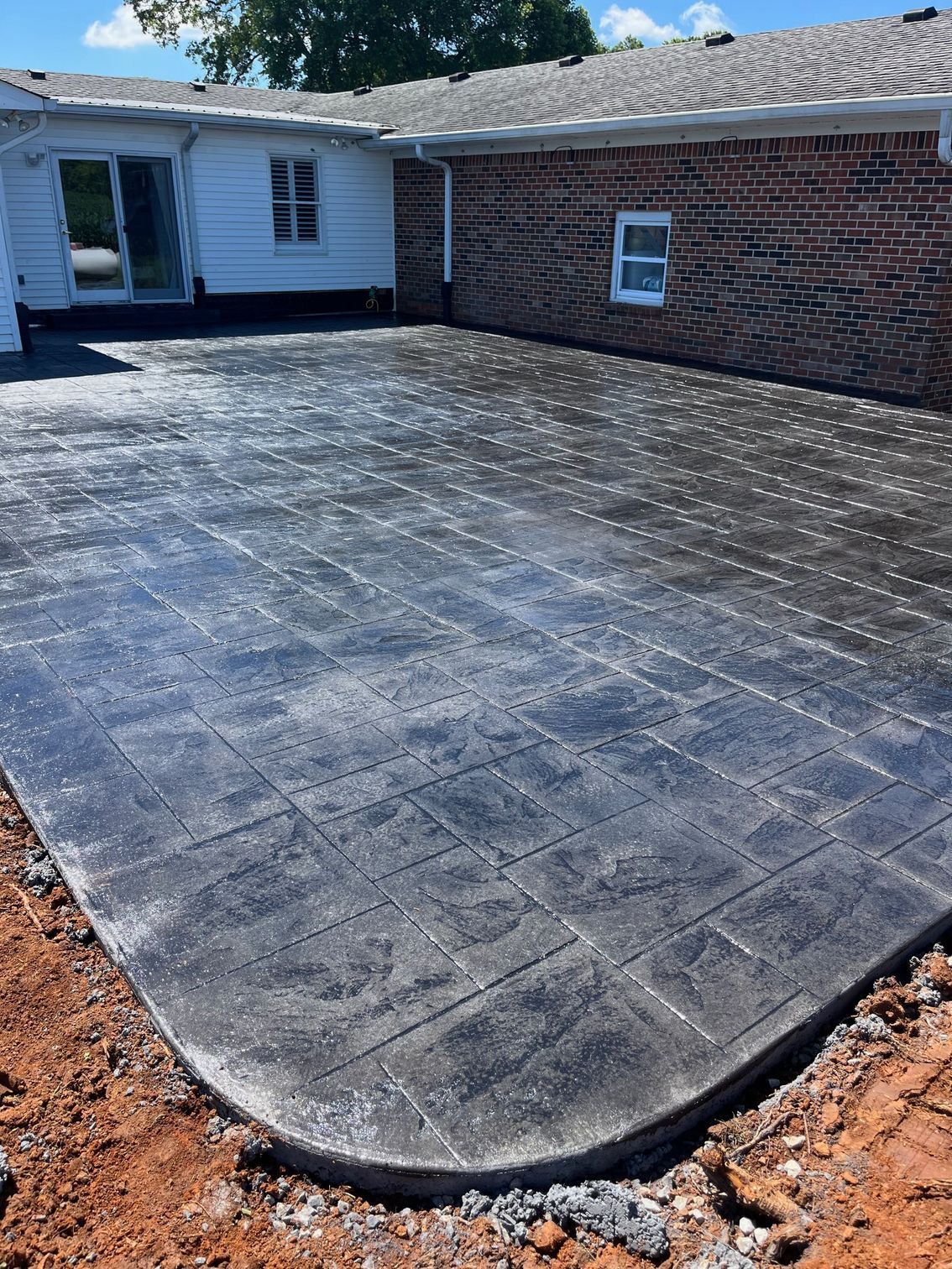 Freshly paved black concrete patio beside a brick house and white siding, with bare dirt edges
