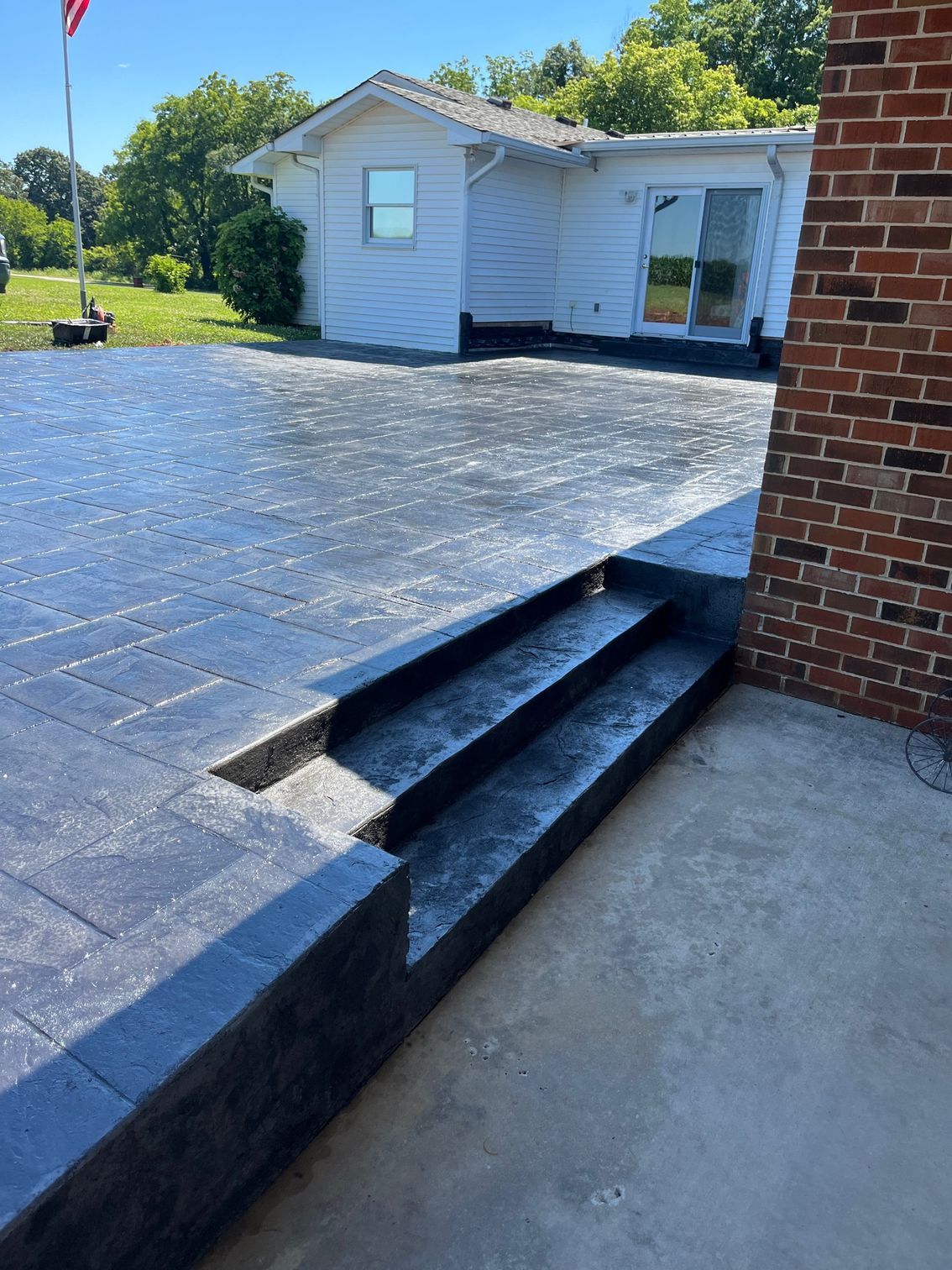 Outdoor patio with dark pavers, steps, and a brick wall beside a white house and lawn