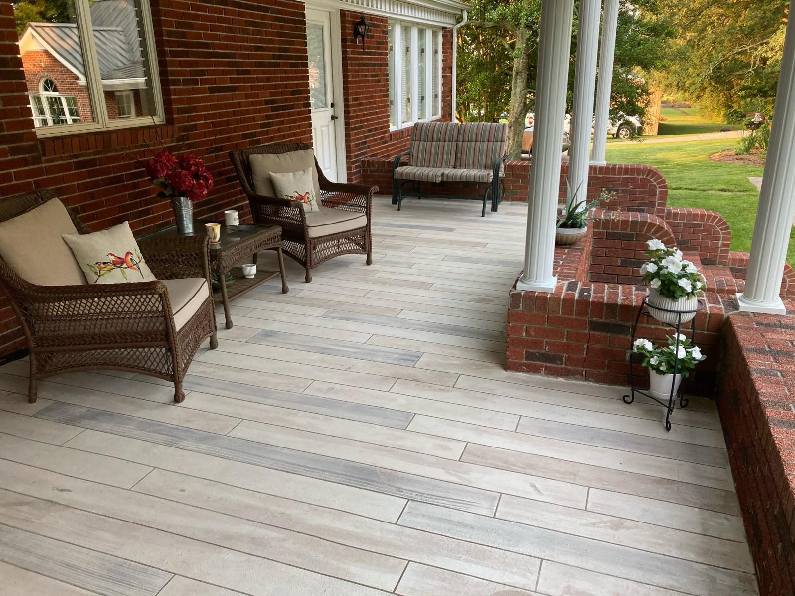 Front porch with wicker chairs, white columns, potted flowers, and a pale wood deck beside a brick house.