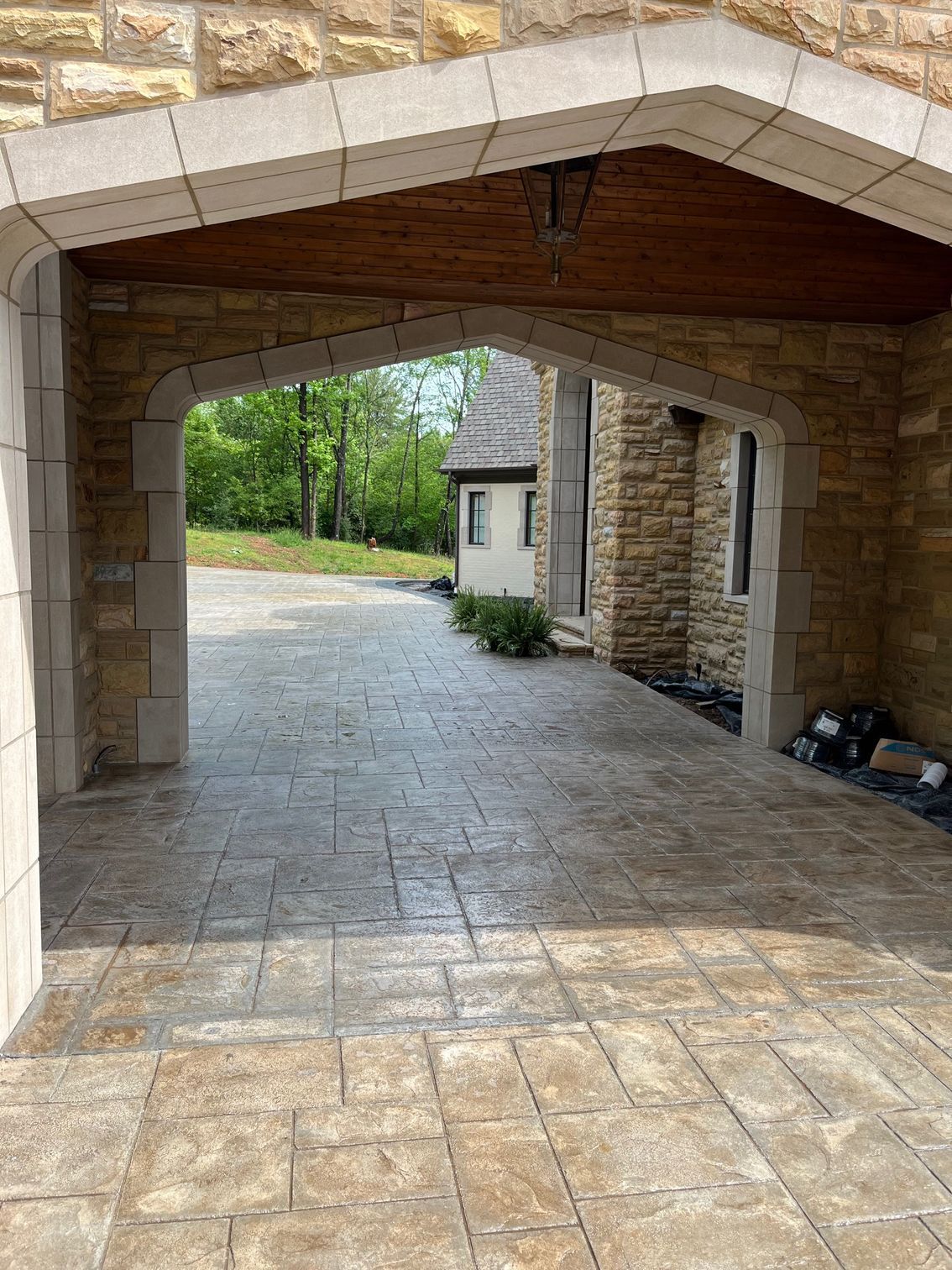 Stone archway opening onto a sunlit paved courtyard with a small building and greenery beyond