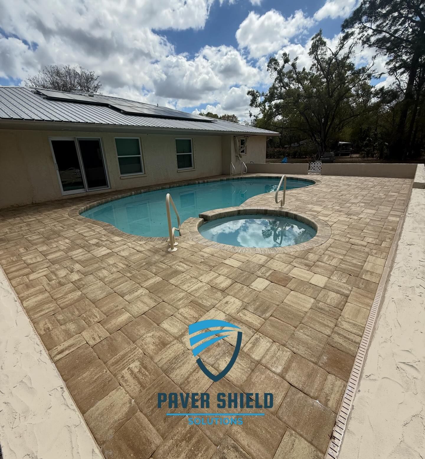 Backyard pool with small spa, tan paver deck, and a house under a cloudy sky