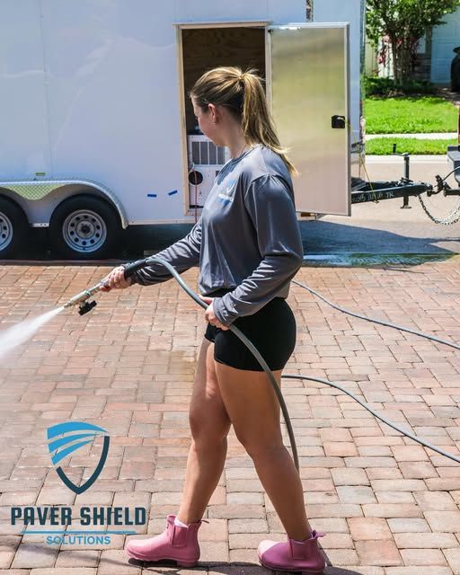 Woman in gray shirt and black shorts walking a leash on a brick driveway beside a trailer