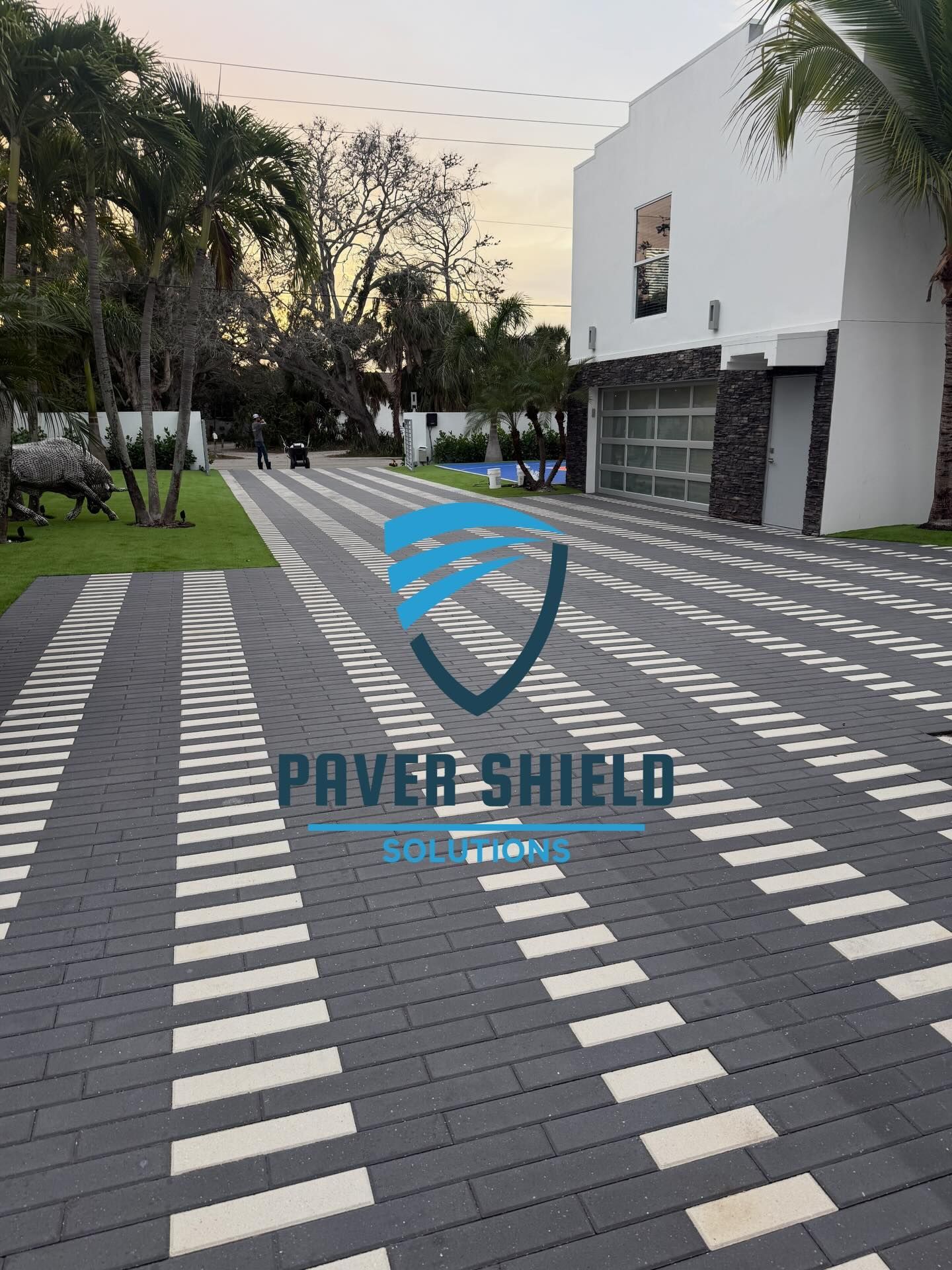 Patterned brick walkway beside a white building and trees, with a Pavishield logo on the pavement.