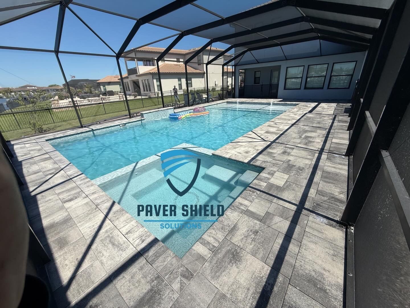 Screened-in backyard pool with gray pavers and a tan house under a clear blue sky