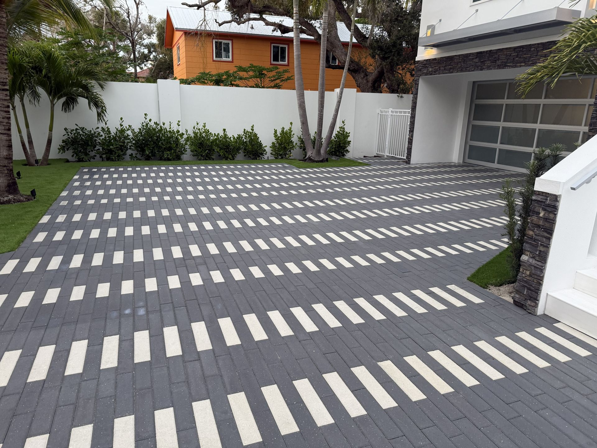 Modern driveway with gray-and-white striped pavers, bordered by white walls and greenery