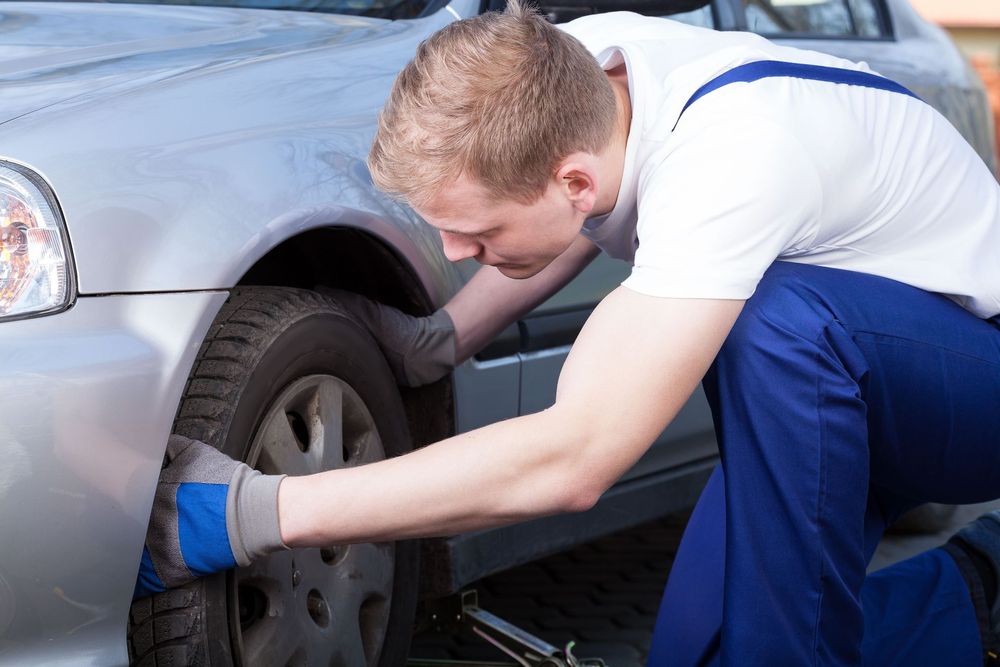 Mechanic in blue overalls and gloves inspecting car tire, kneeling beside a silver car.
