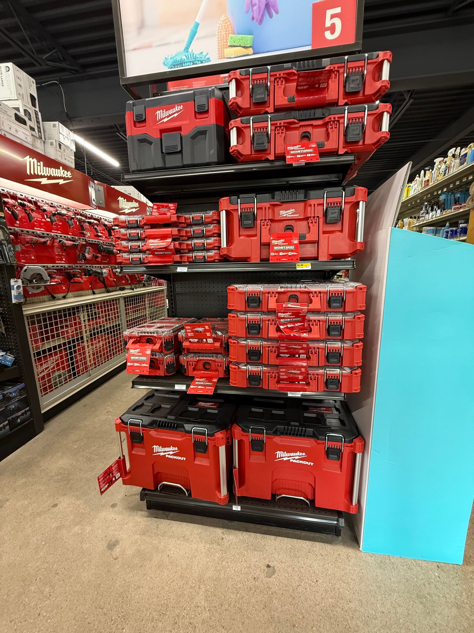 Shelves stocked with red Milwaukee toolboxes in a store. A blue informational board is on the right.