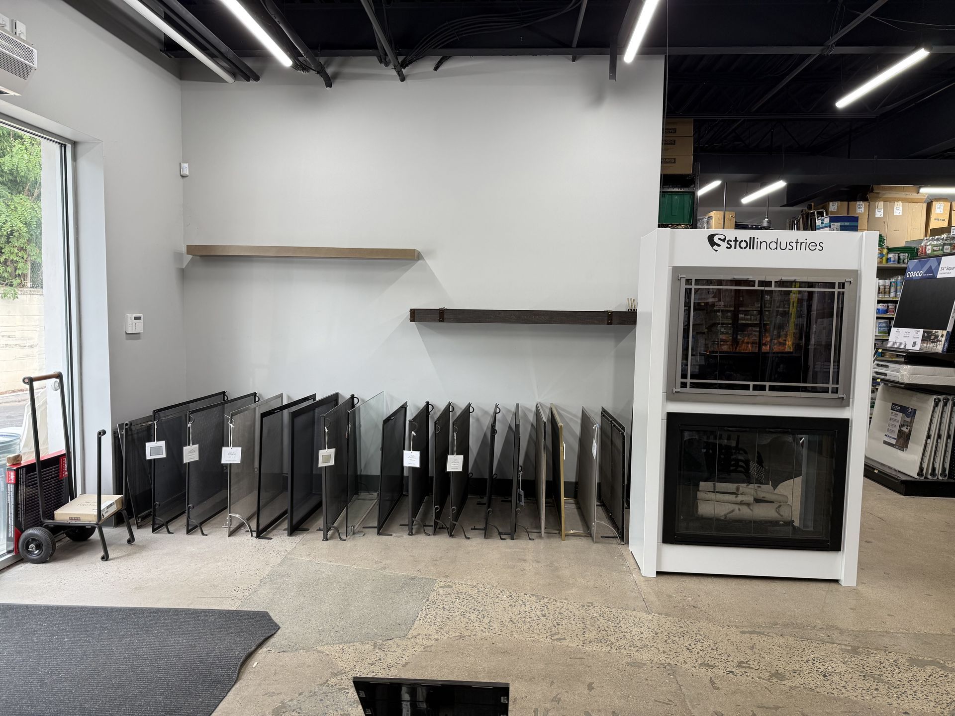 Interior of a store displaying glass panels and a fireplace model on a white stand against a white wall.