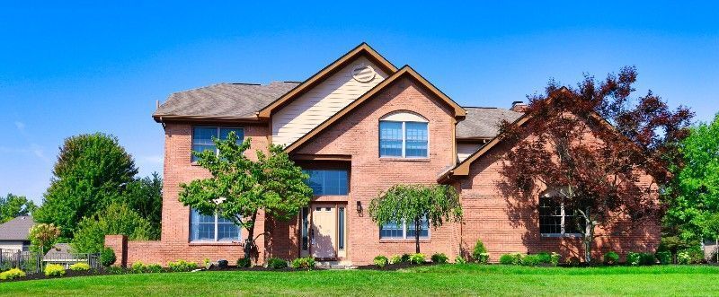 Red brick house with a brown roof and a blue sky in the background. Green trees and grass in front.