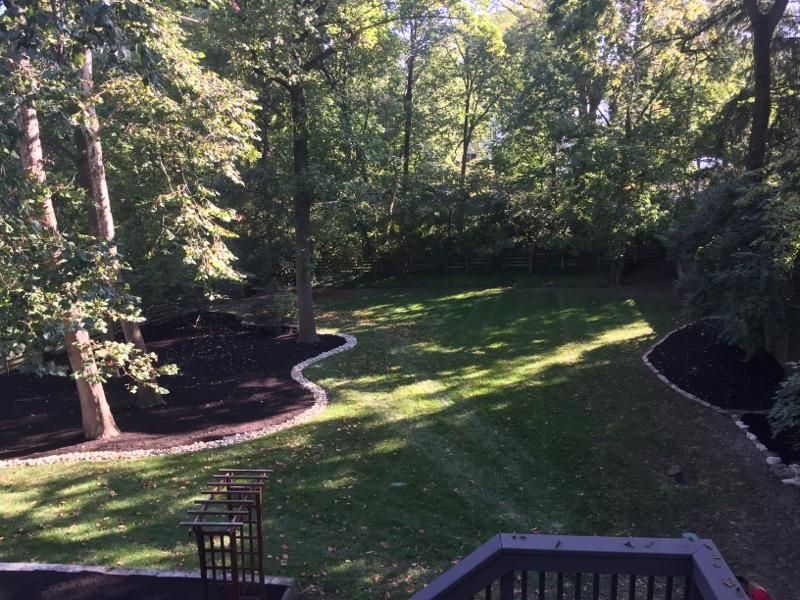 Lush green backyard with trees, manicured lawn, dark mulch borders, and a glimpse of a deck railing.