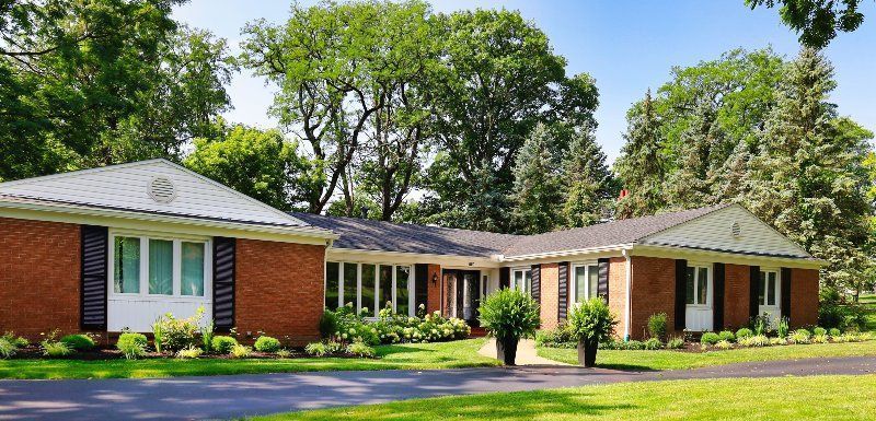 Brick house with dark shutters, white trim, and green lawn. Lush trees and blue sky backdrop.