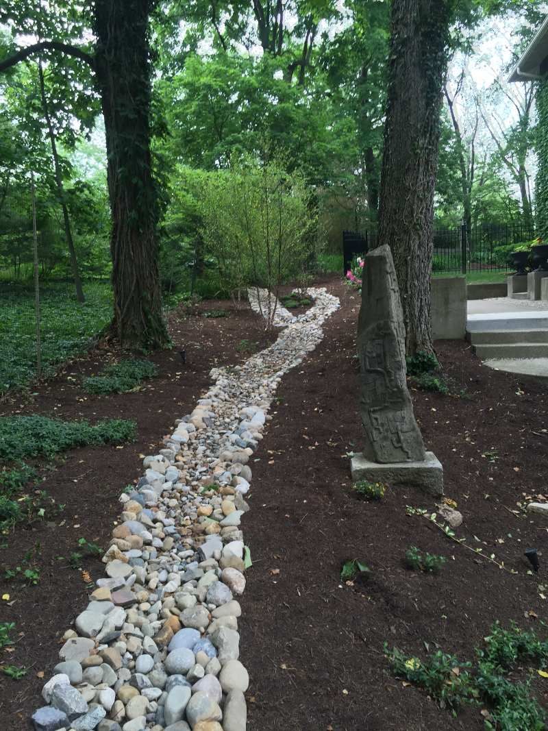 A rock-lined path winds through a wooded area with trees, mulch, and greenery.
