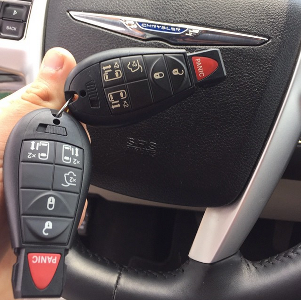 Hand holding two black car key fobs with red panic buttons and silver emblems.