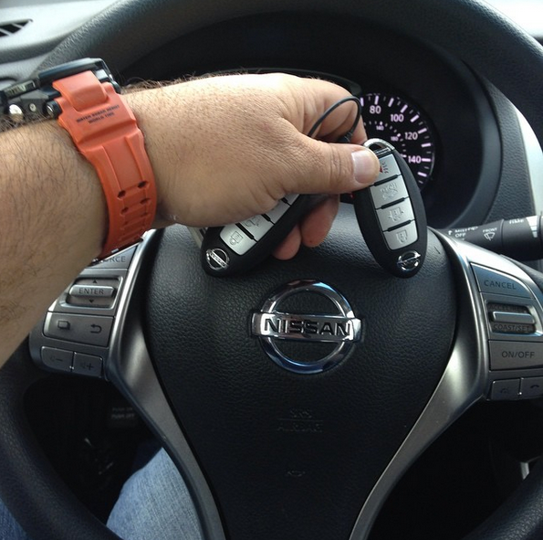 Hand gripping car keys over a Nissan steering wheel, with orange watch strap visible.