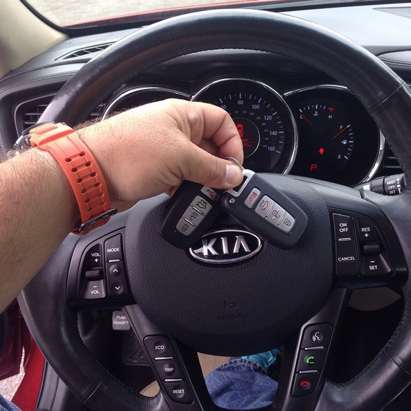 Hand holding Kia key fob over steering wheel inside a car.