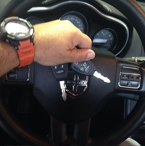 Hand on car steering wheel with key fob and orange watch, dashboard gauges visible