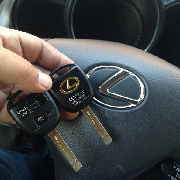 Hand holding two Lexus car keys in front of a Lexus steering wheel logo