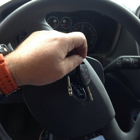 Hand holding a car key in the ignition, with a steering wheel and dashboard visible.