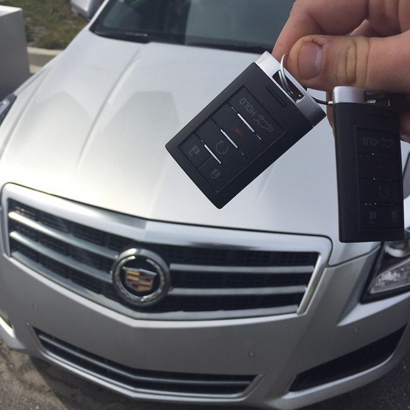 Hand holding two car key fobs in front of a silver Cadillac grille