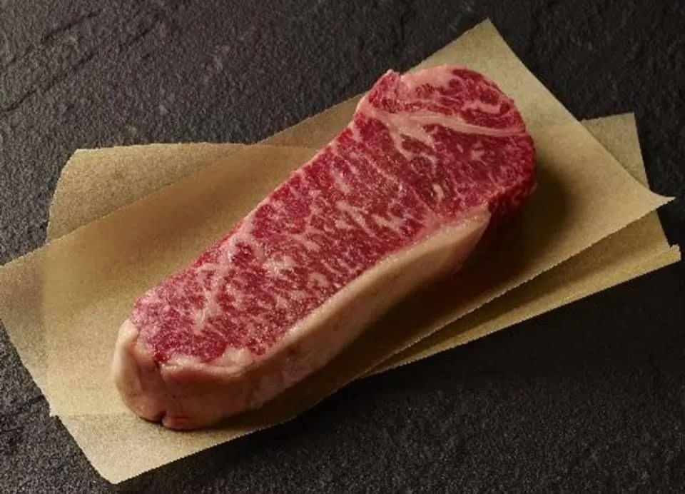 Strip steak with marbled fat on parchment paper, dark background.