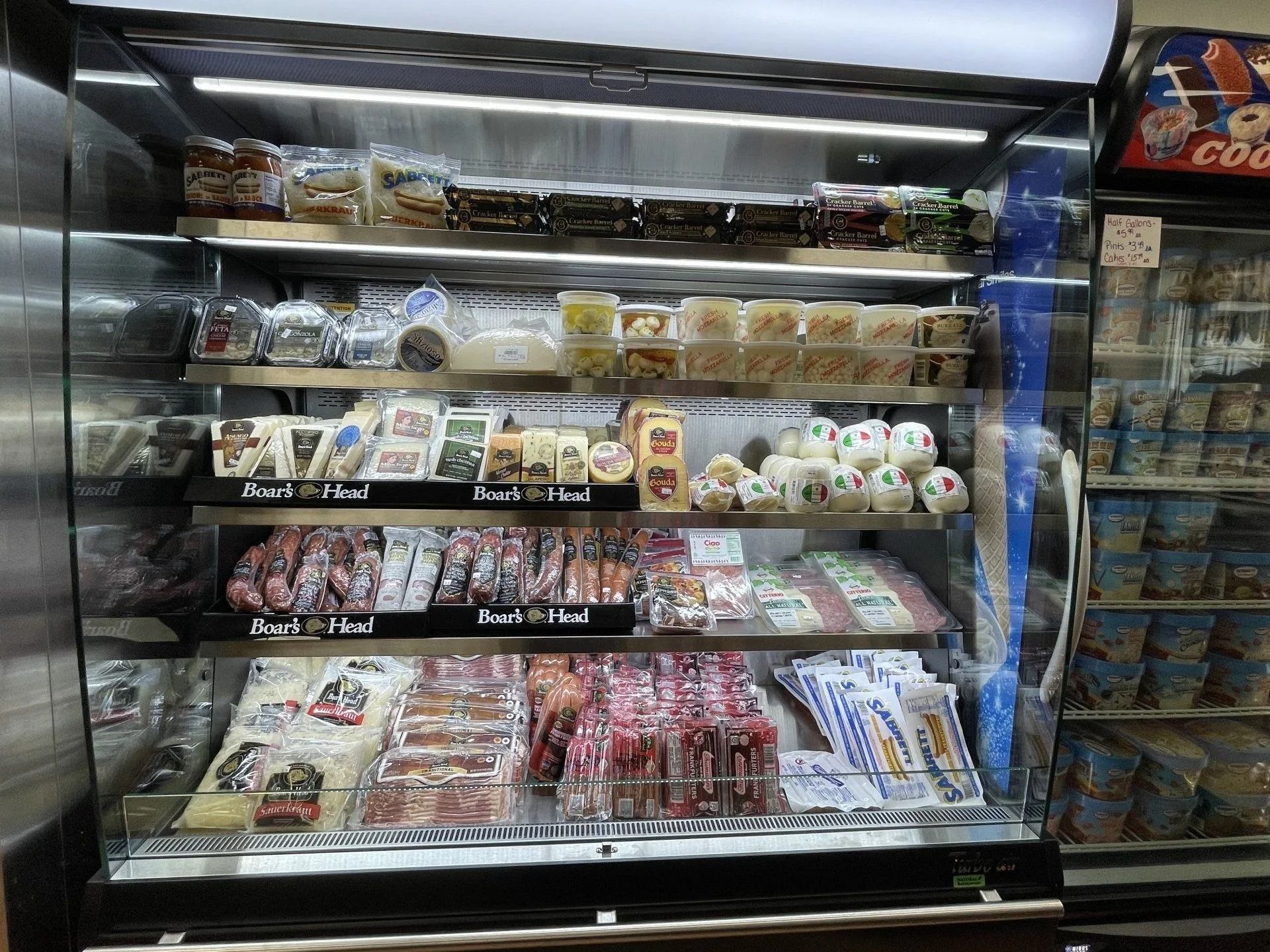 Refrigerated display case with packaged food items: cheese, meats, desserts.
