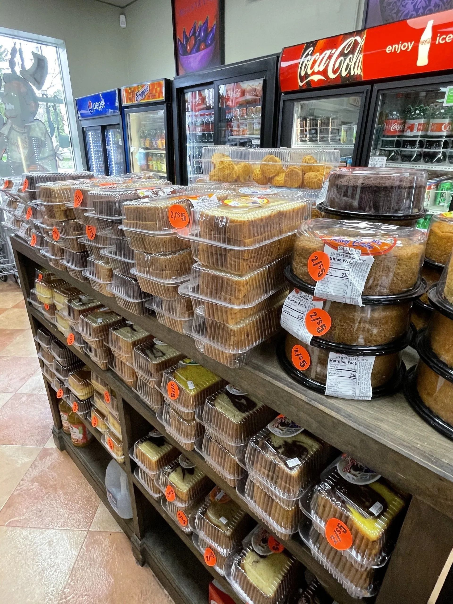 Shelves overflowing with various packaged cakes and pastries in a store setting, with drink coolers visible.