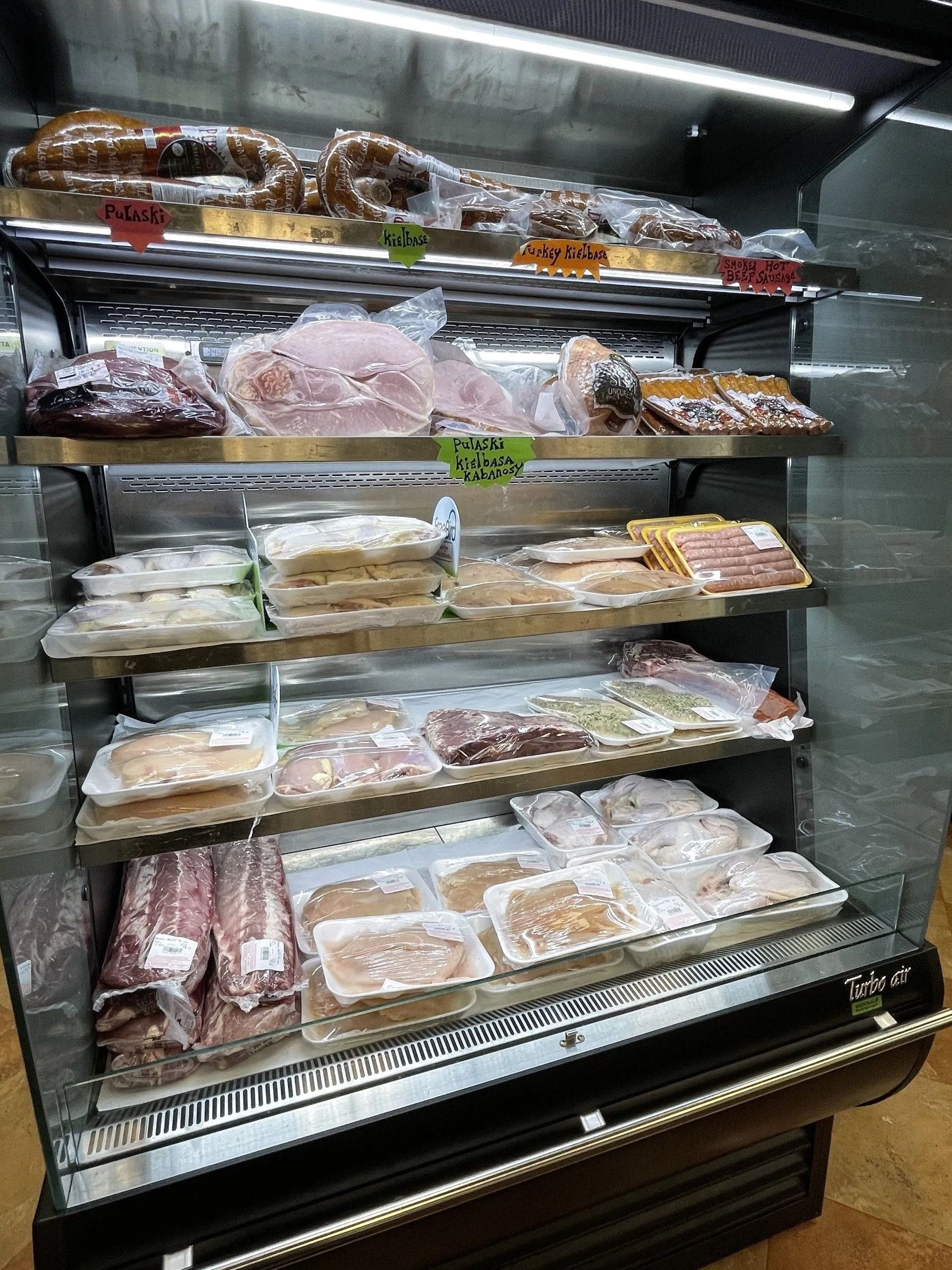 Refrigerator case displaying various vacuum-sealed meats: sausages, ham, bacon, and other cuts.