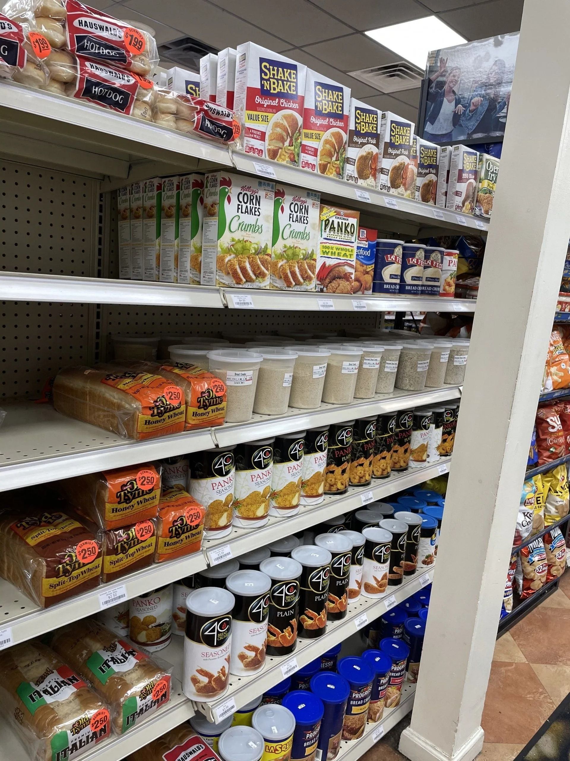 Shelves stocked with packaged food items in a store. Bread, canned goods, jars, and other groceries are visible.