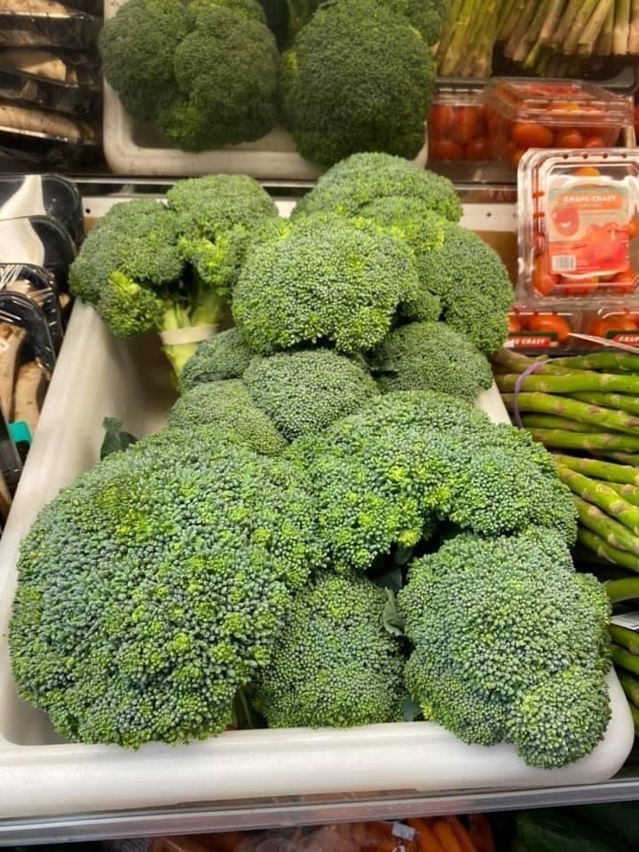Pile of fresh green broccoli florets in a white bin, with asparagus and cherry tomatoes visible.