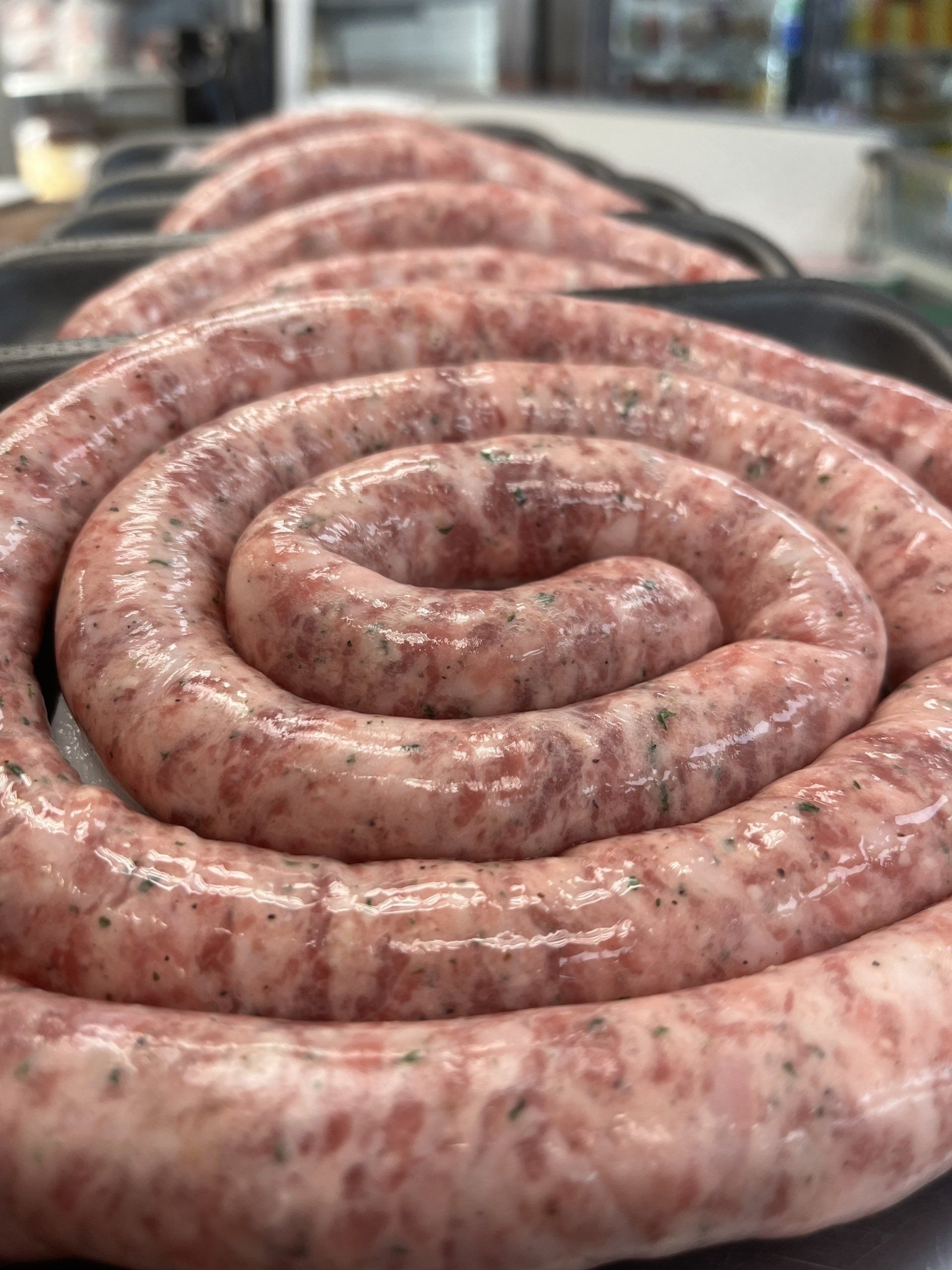 Coiled raw sausage in a black tray, close-up. Pale pink meat with herbs, in a butcher shop setting.