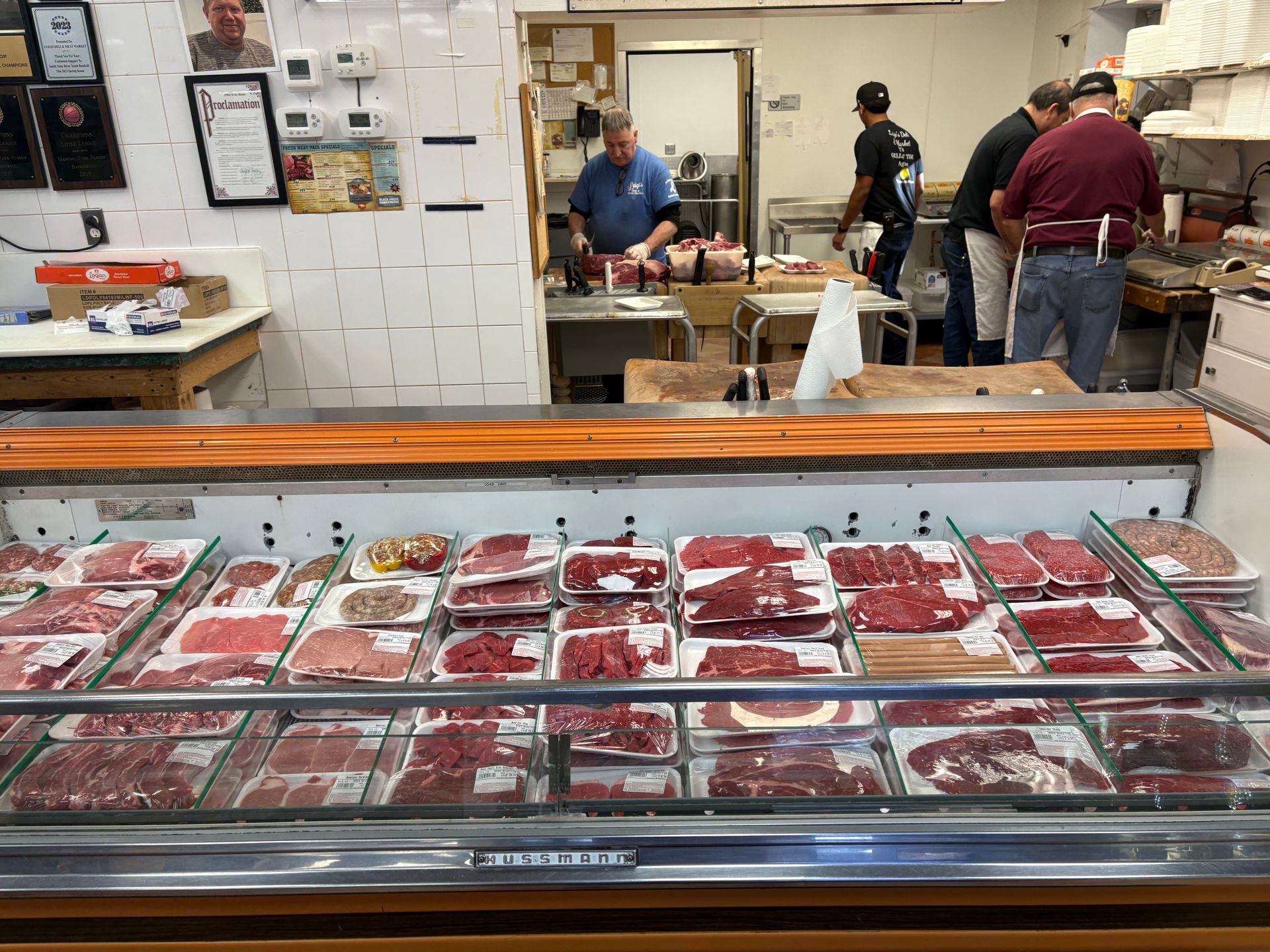 Meat display case in a butcher shop, workers behind the counter preparing cuts.